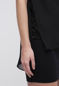 Indumento nero a strati con una sovrapposizione trasparente, tessuto interno texturizzato e orlo liscio. Primo piano di una mano appoggiata sul bordo.