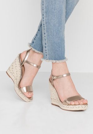 Metaliczne brązowe sandały na koturnie z paskowym wzorem, posiadające teksturowaną, plecioną platformę oraz zapięcie na kostce. Noszone z przetartymi niebieskimi dżinsami.