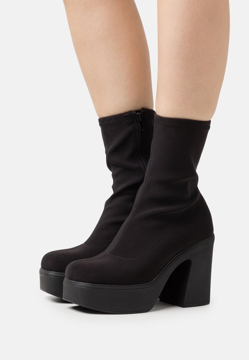 Bottines noires à talons hauts avec semelles plateformes portées sur jambes nues, avec fermetures éclairs latérales et dessus en tissu extensible.