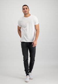 Bílé tričko s krátkým rukávem a jemným zlatým potiskem, v kombinaci s tmavými džíny a bílými teniskami. Jednoduchý, neformální outfit s čistými liniemi.