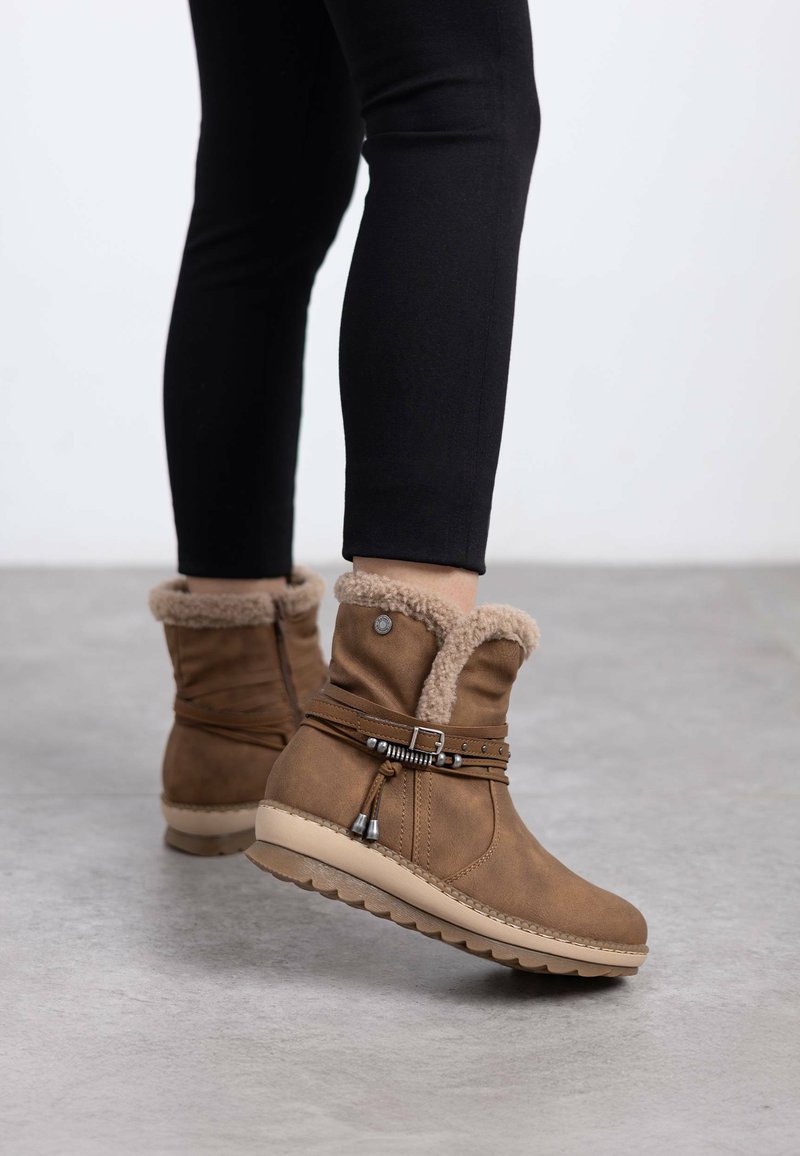 Botas de tobillo marrón con un suave cuello de piel sintética, correa decorativa con acentos metálicos y una suela de goma texturizada.
