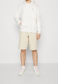 Persona vestida con sudadera con capucha color blanco sucio, pantalones cortos beige hasta la rodilla y zapatillas altas blancas con cintas rosas y el swoosh verde de Nike.