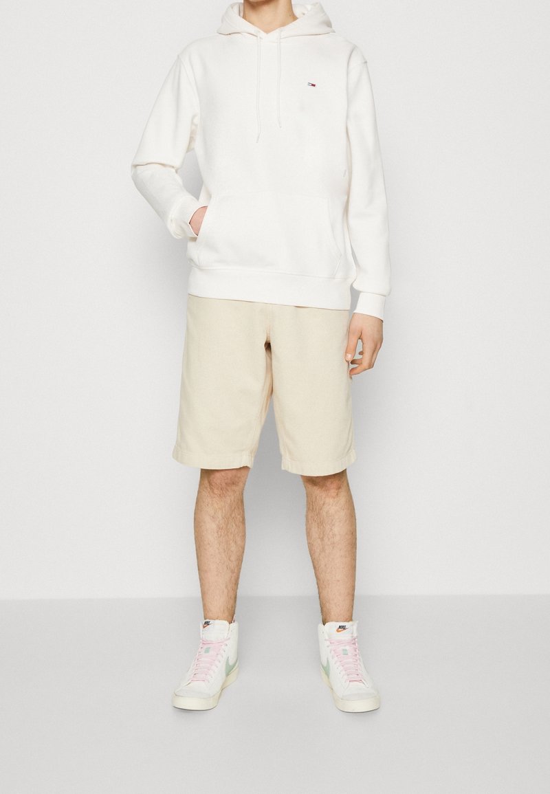 Persona vestida con sudadera con capucha color blanco sucio, pantalones cortos beige hasta la rodilla y zapatillas altas blancas con cintas rosas y el swoosh verde de Nike.