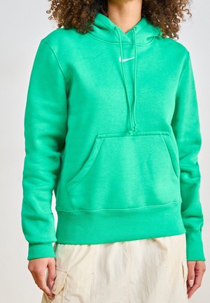 Personne portant un sweat à capuche vert vif avec un petit logo Nike blanc et une jupe beige, debout devant un fond uni.