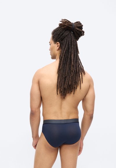 Άνδρας με μακριά dreadlocks δεμένα πίσω, φορώντας σκούρα μπλε εσώρουχα, στέκεται με την πλάτη προς τα εμπρός σε ένα απλό φωτεινό φόντο.