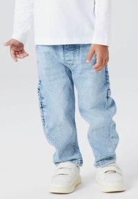 Šviesiai mėlyni džinsai su laisvu kirpimu, turintys šoninius cargo kišenes. Apsirengti su baltais sportbačiais, kuriuos laiko dvi Velcro juostos.