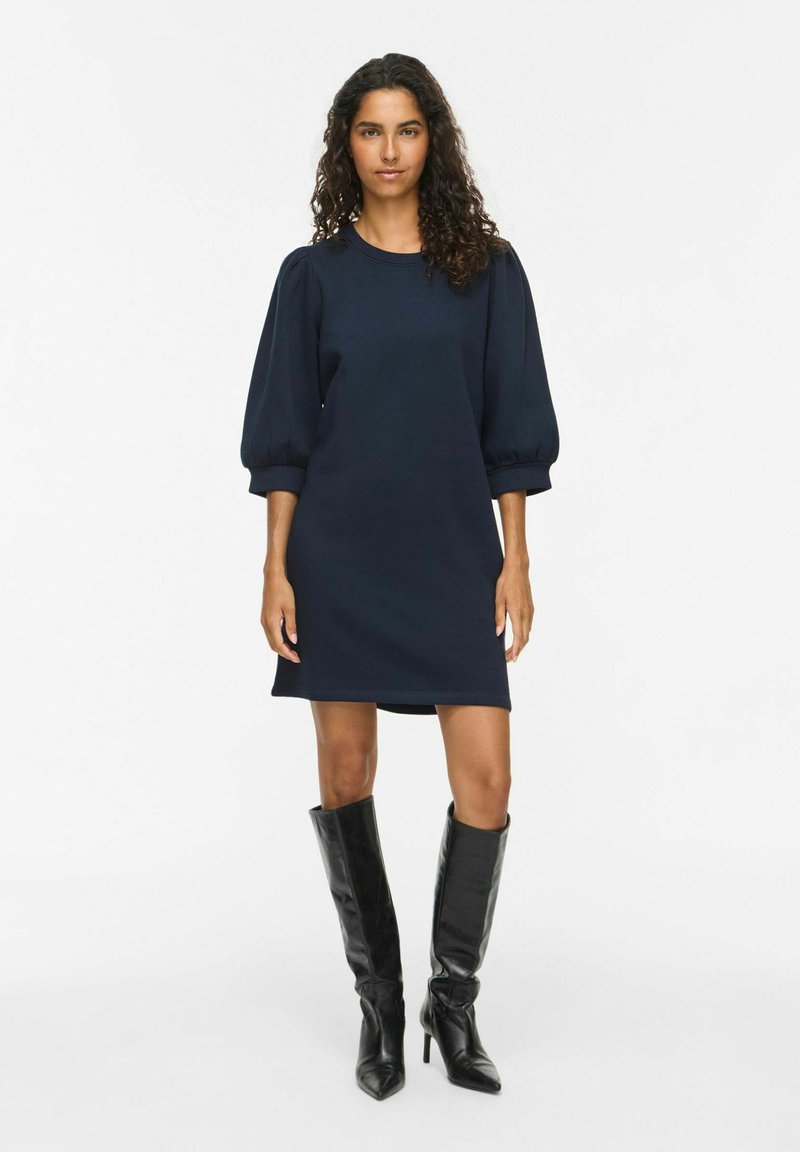 Femme aux cheveux bouclés portant une robe bleu marine avec des manches ballons et des bottes noires montantes à talons, debout sur un fond blanc.