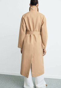 Palton bej cu guler înalt și curea, având o despicătură în spate. Fabricat din material neted, cu un design până la genunchi.