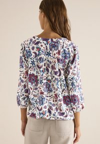 Femme aux longs cheveux bruns portant un chemisier blanc à motifs floraux bleus et rouges et un pantalon beige, debout devant un fond uni.