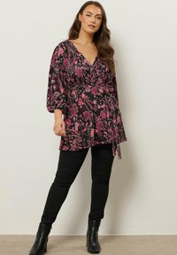 Top nero avvolgente con motivo floreale rosa, maniche lunghe e cintura in vita, abbinato a pantaloni skinny neri e stivaletti.