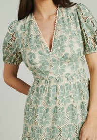 Mujer vistiendo un vestido de manga corta con un superposición de encaje floral verde claro y escote en V profundo, frente a un fondo liso.
