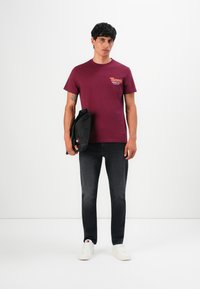 Mann trägt ein bordeauxfarbenes Tommy Jeans T-Shirt, schwarze Jeans, weiße Sneaker und hält eine schwarze Tasche vor einem schlichten weißen Hintergrund.