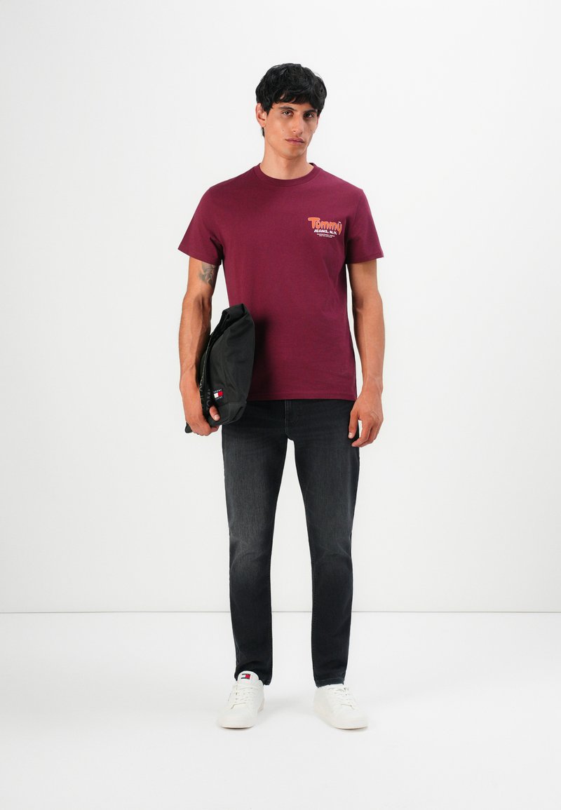 Man wearing burgundy Tommy Jeans t-shirt, black jeans, white sneakers, holding a black bag against a plain white background.