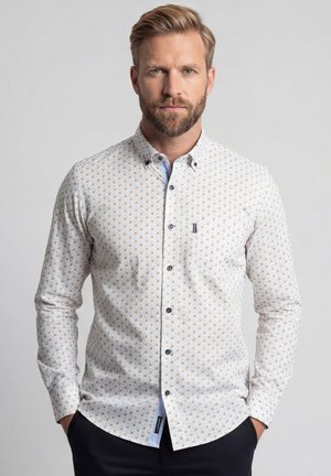 Blonde man met baard, draagt een wit overhemd met een klein bruin polkadotpatroon, handen in de zakken, neutrale achtergrond.