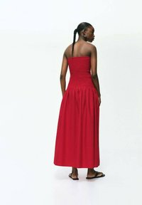Robe maxi rouge sans manches avec un corsage froncé et une jupe fluide. Le tissu semble doux avec une texture lisse. Portée avec des sandales plates.