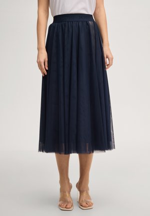 Femme portant une jupe midi en tulle bleu marine avec une taille froncée et un top blanc sans manches, debout pieds nus dans des sandales transparentes à brides.