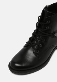 Bottine noire en cuir à lacets avec bout arrondi et œillets métalliques sur fond blanc.