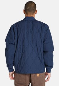 Marinblå quiltad jacka med ett texturerat mönster, ribbat krage och ärmslut, samt en liten märkespatch i nedre delen av ryggen.