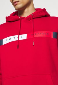 Rode pullover hoodie van katoen, met een witte en navy horizontale streep waarop "TOMMY HILFIGER" in het zwart is gedrukt. Inclusief drawstrings met metalen punten.