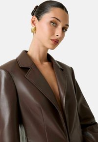 Blazer in pelle marrone con rever a incavo, scollo profondo e finitura strutturata; orecchini geometrici dorati completano il look.