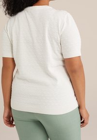 Biały sweter z teksturowanym wzorem, krótkim rękawem, z prążkowanym dekoltem i krawędzią, zestawiony z jasnymi, dopasowanymi zielonymi spodniami, ukazujący gładkie wykończenie tkaniny.