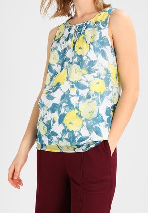 Mouwloze witte blouse met geel en blauw bloemenprint, gedragen met bordeauxrode broek, linkerhand van het model in de zak tegen een effen achtergrond.