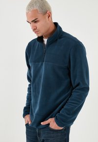 Pullover in pile blu navy con colletto a mezza zip, tessuto morbido e pannello sul petto a contrasto. Il modello lo abbina a jeans in denim scuri.