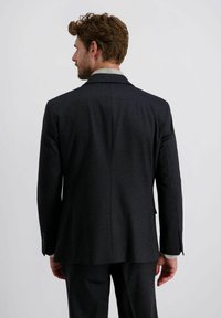 Man met bruin haar en baard, gekleed in een donker getextureerd pak, staande met zijn gezicht naar weg gericht tegen een effen lichte achtergrond.