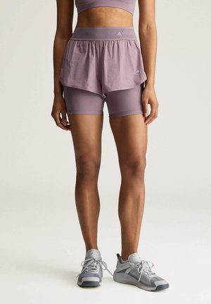 Vrouw die mauve tweedelige sportshorts en een bijpassende sportbeha draagt, blootvoets staand op een lichte achtergrond.