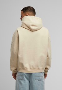 Cremefarget hettegenser laget av myk stoff, med en avslappet passform og sidelommer. Bakfra viser den et minimalistisk design uten trykk.