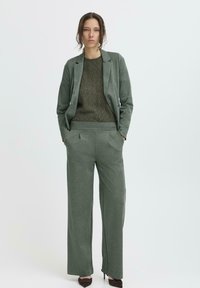Sacou verde croit și pantaloni largi asortați, dintr-un material moale, asociați cu un pulover texturat de culoare verde închis.
