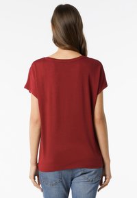 T-shirt rosso a maniche corte con una vestibilità rilassata e un ampio scollo. Tessuto morbido e liscio con un design semplice. Vista posteriore che mostra cuciture piatte.