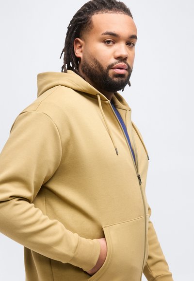 Άνδρας με γένια και κότσους, ντυμένος με ταμπά φούτερ με φερμουάρ και τα χέρια στις τσέπες, κοιτάζει προς την κάμερα πάνω σε ένα απλό φωτεινό φόντο.