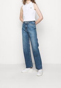 Biała bezrękawnikowa koszulka polo z kołnierzykiem, zestawiona z niebieskimi dżinsami o wysokim stanie i prostych nogawkach. Noszona z białymi trampkami.