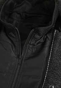 Czarna kurtka zapinana na zamek, charakteryzująca się teksturowanym zewnętrznym materiałem oraz gładką podszewką, z precyzyjnymi przeszyciami i małą metką przy zamku.