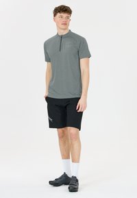 Grå kortärmad zip-hals tröja, texturerad tyg. Matchas med svarta shorts med en logga. Modellen bär svarta cykelskor och vita strumpor.