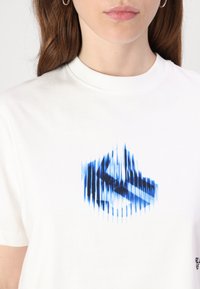 Bela bombažna majica z modrim grafičnim oblikovanjem in vertikalnimi črtami ter stilizirano logotipom na sredini. Kratke rokave in okroglo izrezovanje.