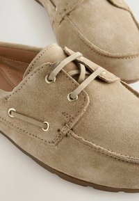 Beige suede boat shoes with round toe, decorative lacing, gold-tone eyelets, and minimal stitching details. Soft texture and flat sole.