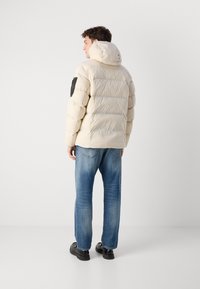 Jachetă puffer bej cu glugă, cu cusături orizontale, un patch negru pe brațul stâng, purtată cu blugi albaștri și pantofi negri.