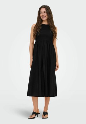 Femme aux longs cheveux bouclés portant une robe midi noire sans manches et des sandales à talons noires, souriante, debout sur un fond uni.