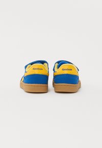 Zapatillas azules y amarillas con textura de ante, que cuentan con una correa de Velcro, un collar acolchado y una suela de goma marrón. Logo de Reebok en el talón.
