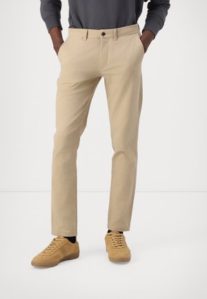 Beige Chinos mit schmaler Passform, aus Baumwolle, mit Vordertaschen und schwarzem Knopfverschluss, kombiniert mit hellbraunen Sneakers.