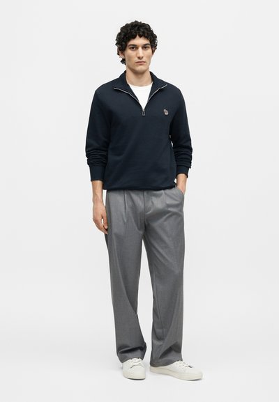 Νέος άνδρας που στέκεται με το ένα χέρι στην τσέπη, φοράει μπλε πουλόβερ με φερμουάρ μέχρι το στήθος, γκρι πλισέ παντελόνι και λευκά αθλητικά παπούτσια.