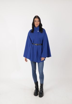 Giacca a mantella blu in tessuto testurizzato, con colletto alto e chiusura a un solo bottone, cinteata con una cintura nera, abbinata a jeans skinny e stivali chunky.