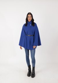 Blå cape-jacka i texturerat tyg, med hög krage och en enda knappstängning, åtdragen med ett svart bälte, kombinerad med slim fit jeans och kraftiga stövlar.