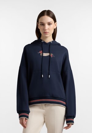 Giovane donna che indossa una felpa con cappuccio blu navy con grafica di bassotto decorata, coulisse e polsini a righe, abbinata a pantaloni beige.