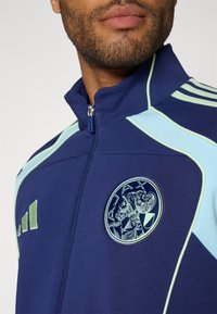 Marineblå sportsjakke med lyseblå detaljer, opretstående krave, lynlåslukning og broderet logo med en klassisk figur.