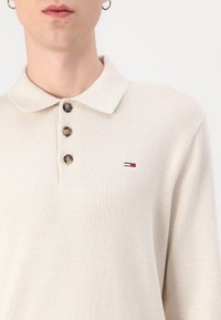 Ljusbeige stickad polo med spridkrage, tre knappar fram och en liten broderad logotyp i röd, vit och blå.