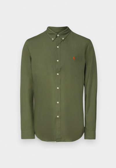 Olīvu zaļa krekls ar garām piedurknēm un pogājamām apkaklēm, ar priekšējo klāju un mazu sarkanu izšūtu logotipu kreisajā krūtī.