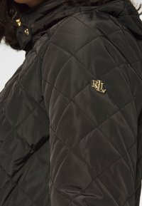 Svart vadderad jacka med en glänsande yta, med en gyllene "RL"-logotyp på axeln och en huva. Tyget har en mjuk textur.